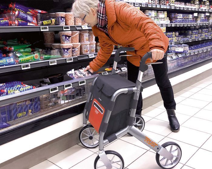 Rollator mit Einkaufstasche. Grauer Rahmen, schwarze Tasche mit roter Front. Person im Supermarkt. "Trust" Schriftzug.
