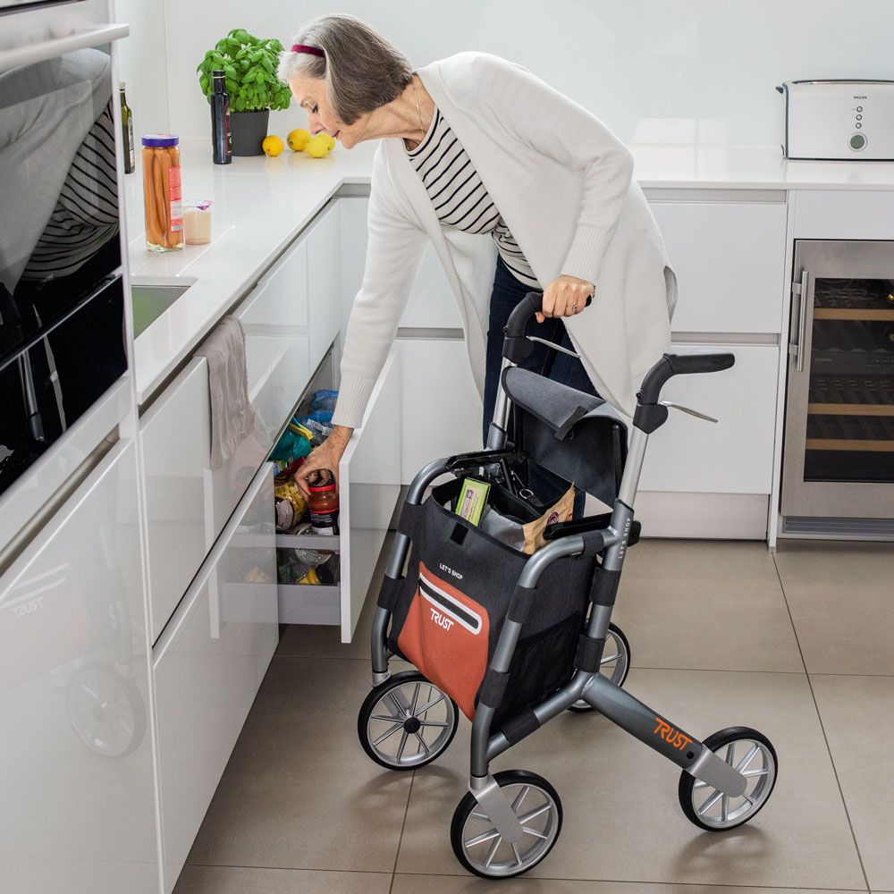 Ältere Frau in Küche mit Rollator. Rollator mit Einkaufstasche, gefüllt mit Lebensmitteln. Grauer Rahmen, orangefarbene Tasche.