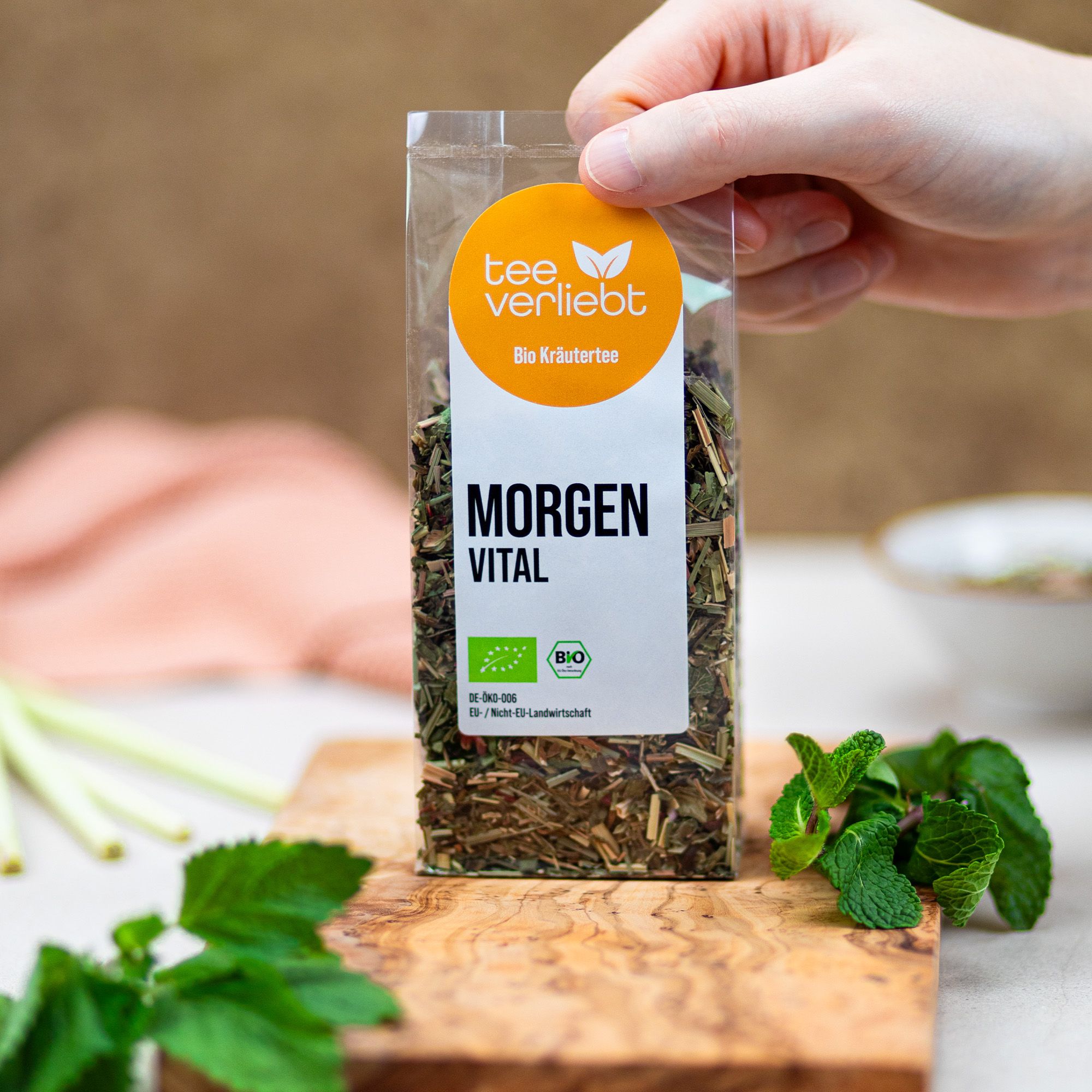 Hand hält Kräutermischung in transparenter Verpackung. Aufdruck: teeverliebt, Morgen Vital, Bio Kräutertee. EU-Bio-Logo. Minze.