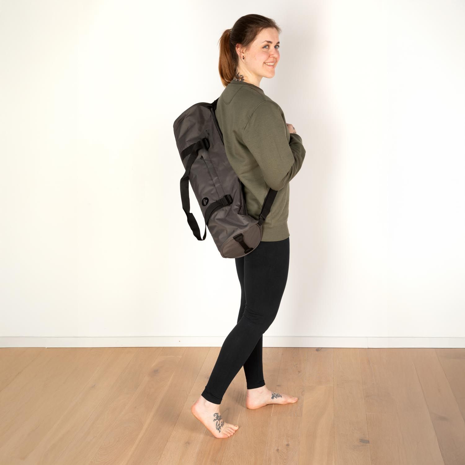 Frau trägt eine graue Sporttasche auf dem Rücken. Schwarze Leggings, grünes Oberteil. Holzfußboden.