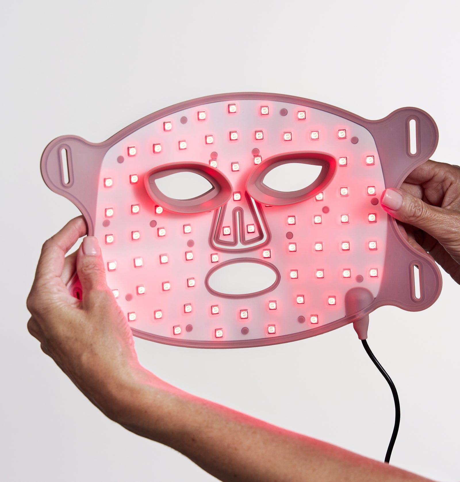 Hände halten eine rosa LED-Gesichtsmaske. Aussparungen für Augen, Nase und Mund. Schwarzes Kabel. Rote LEDs.