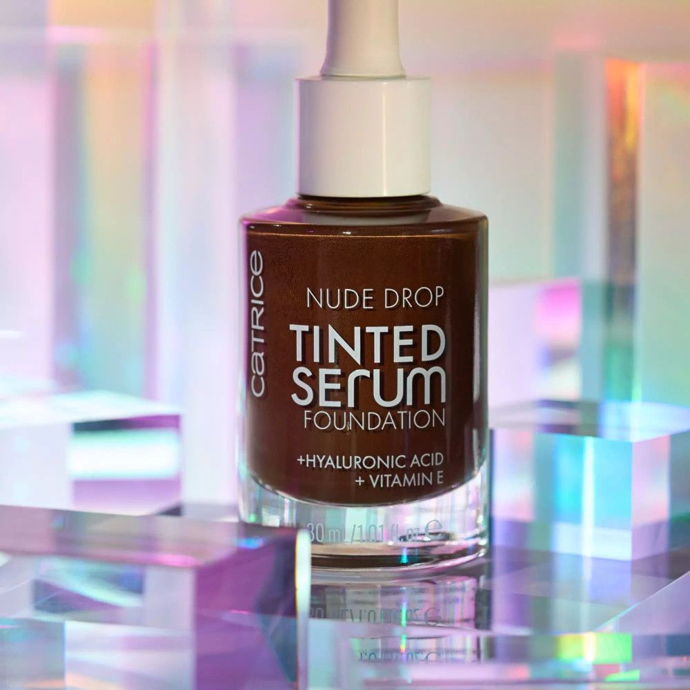 Braune Foundation-Flasche mit Pipette. Aufschrift: Nude Drop Tinted Serum Foundation, + Hyaluronsäure + Vitamin E. 30 ml. Auf glänzendem Hintergrund.