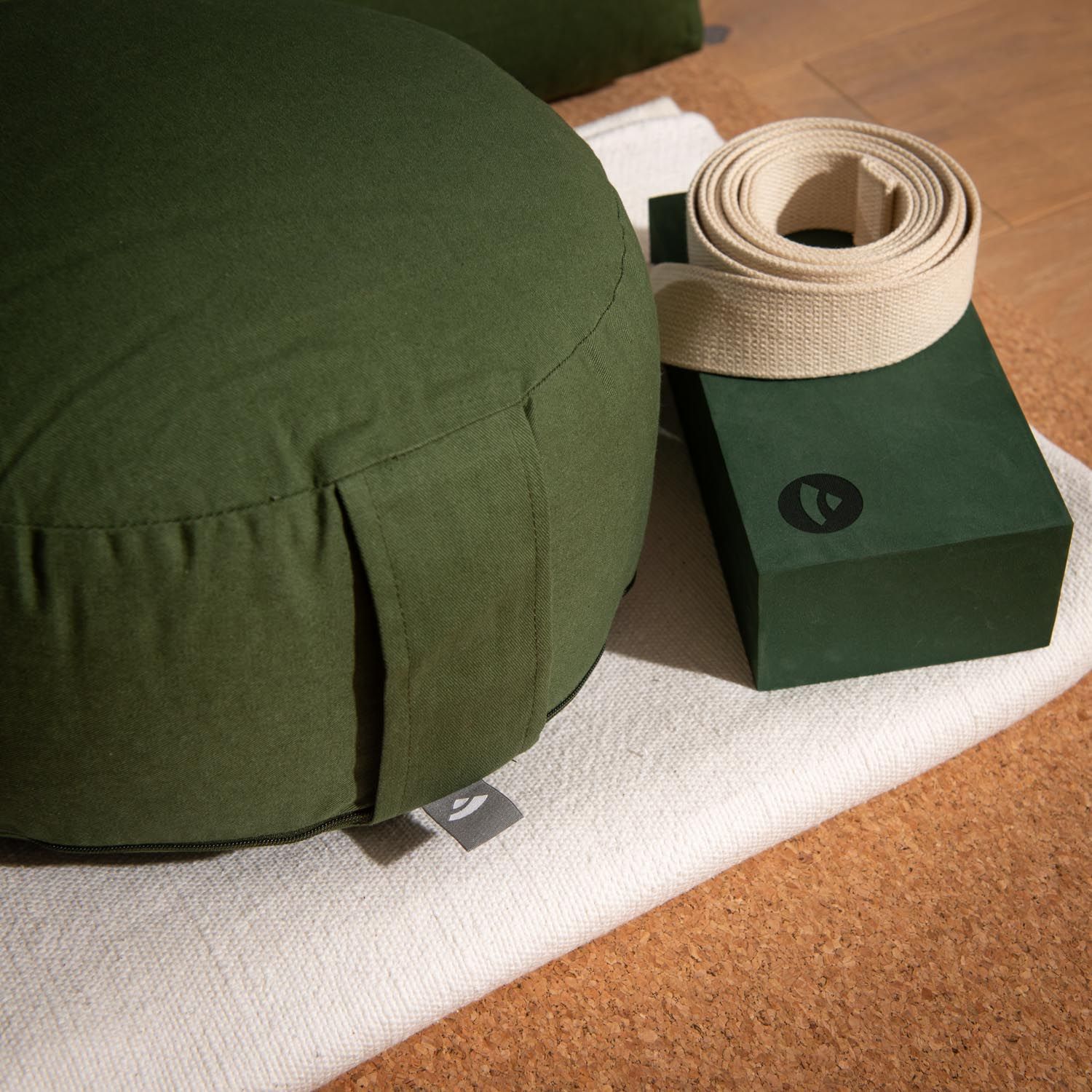 Meditationskissen in Waldgrün neben einem grünen Yogablock mit Yogagurt auf weißem Handtuch.