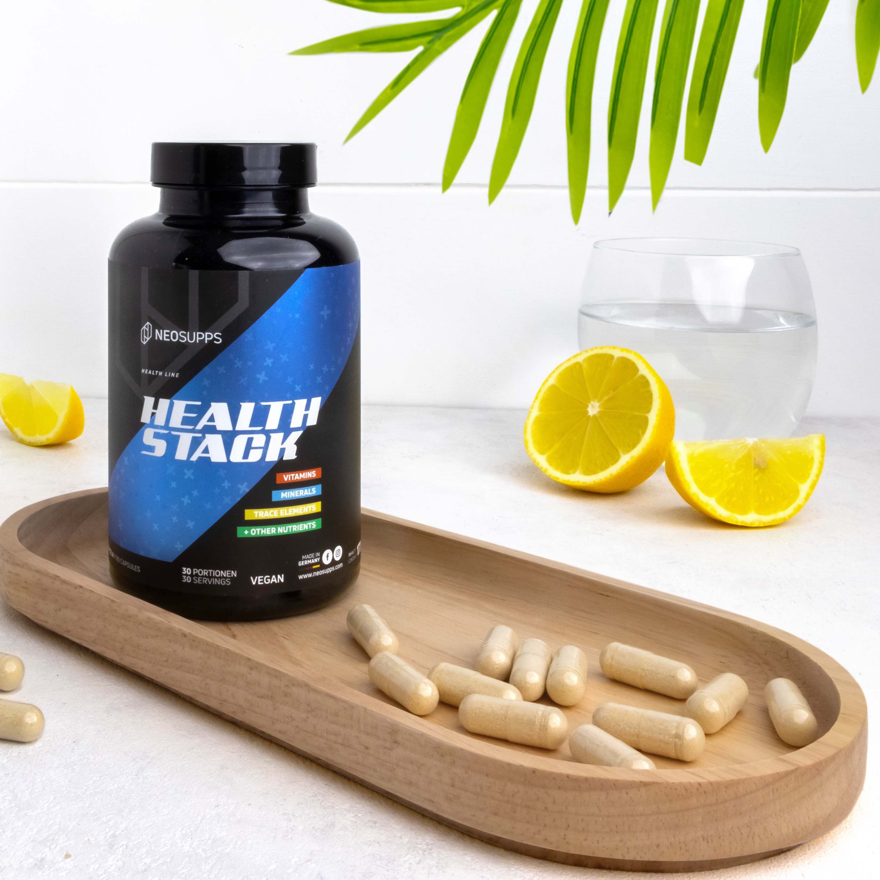 Schwarze Flasche HEALTH STACK auf Holztablett. Daneben Zitronen, Wasserglas und Kapseln. Aufschrift: Neosupps, Health Stack, Vegan.