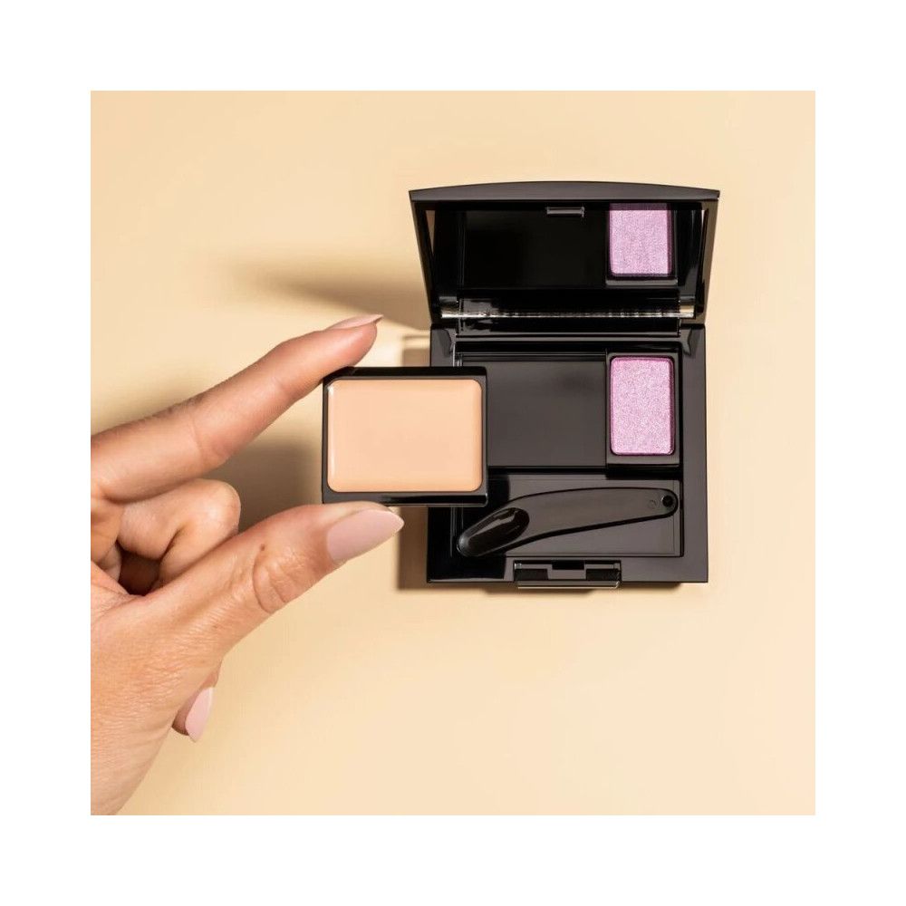 Geöffnete Beauty Box Trio mit Lidschatten und Applikator. Eine Hand hält einen weiteren Lidschatten. Beige Hintergrund.