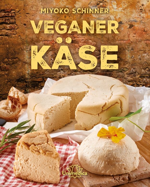 Buchcover mit dem Titel "Veganer Käse". Gezeigt werden verschiedene Käsesorten, teilweise angeschnitten, auf einem Tisch. Im Hintergrund eine Backsteinmauer.