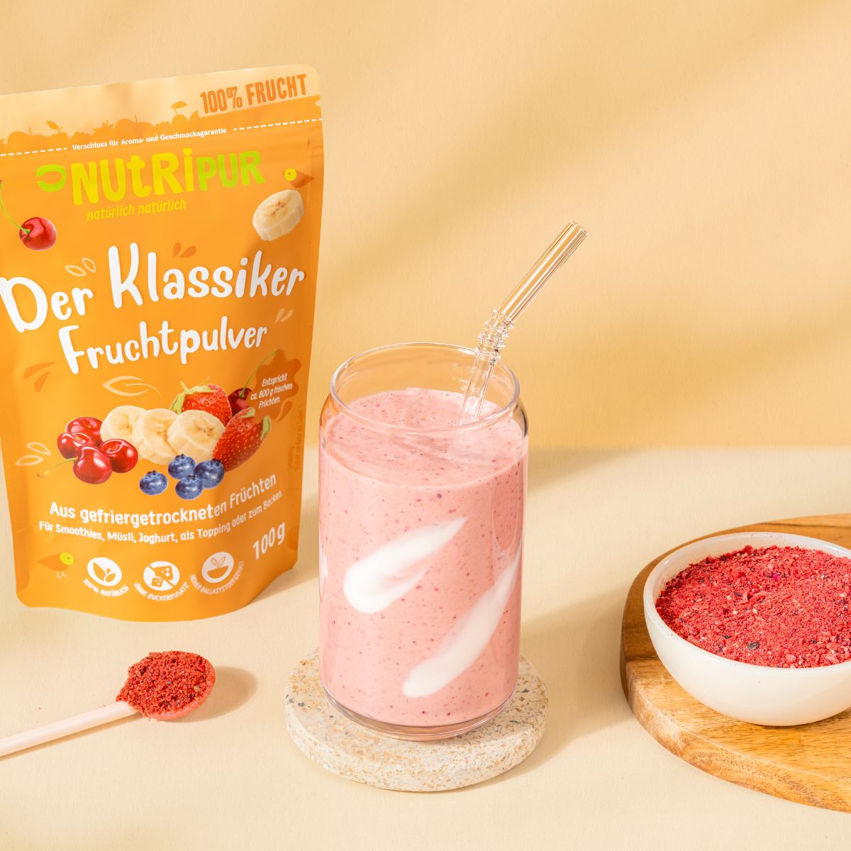Glas mit rosa Smoothie, Strohhalm. Daneben eine Tüte mit Fruchtpulver und eine Schale mit Fruchtpulver. Auf einem Holzbrett.