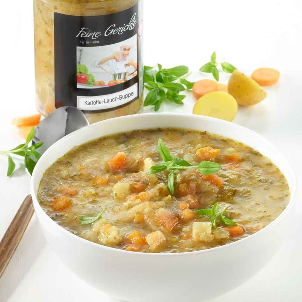 Schüssel mit Schrader Kartoffel-Lauch-Suppe Bio. Gemüse und Kräuter daneben. Glas im Hintergrund.