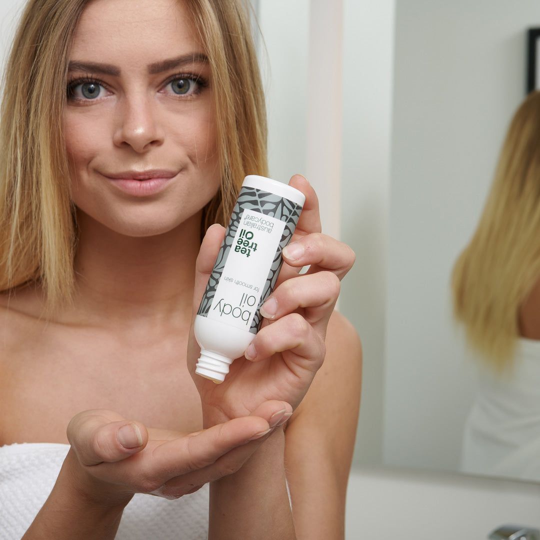 Frau hält eine kleine Flasche Body Oil. Weiße Flasche mit grauen Blättern. Text: Tea Tree Oil, Australian Bodycare.