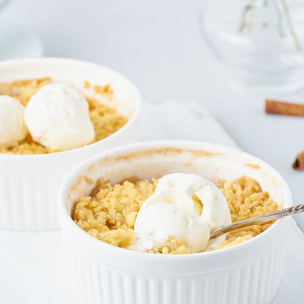 Zwei Schalen mit Apfel-Crumble und Eis. Löffel in einer Schale. Zimtstange im Hintergrund. Helle Umgebung.