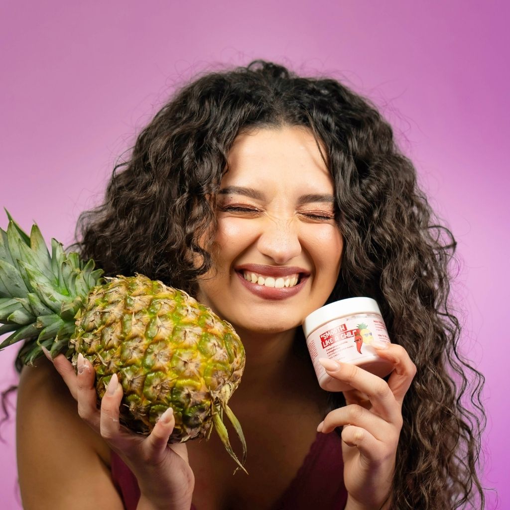 Una donna sorridente tiene in mano un ananas e un barattolo di crema "Smooth Like a Chili". La crema è rosa con un'etichetta che mostra un peperoncino e un ananas. La donna ha i capelli ricci e indossa un top rosso.