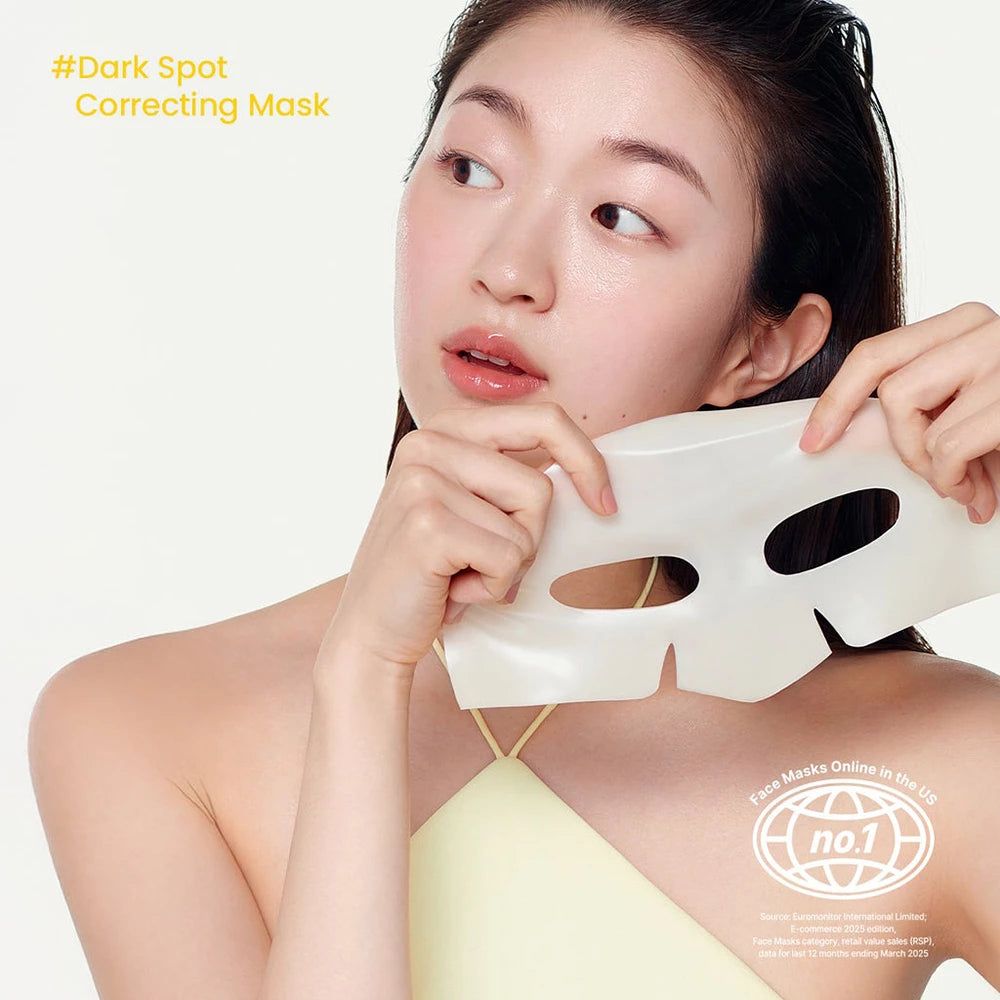 Person hält Gesichtsmaske. Weiße Maske mit Aussparungen für Augen und Mund. Gelbes Oberteil, Text im Hintergrund.