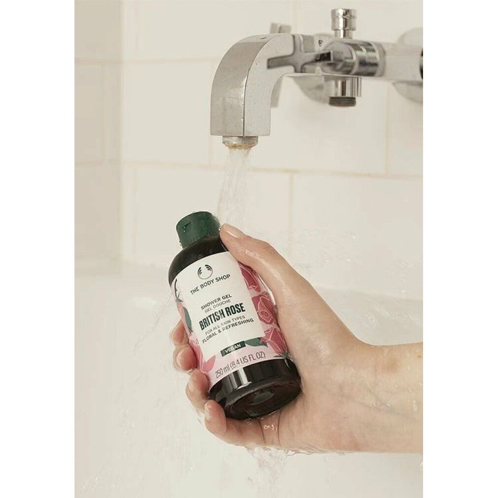 Hand hält Duschgel-Flasche British Rose unter Wasserstrahl. Grüne Kappe, weißes Etikett mit Rosenmuster. Vegan.