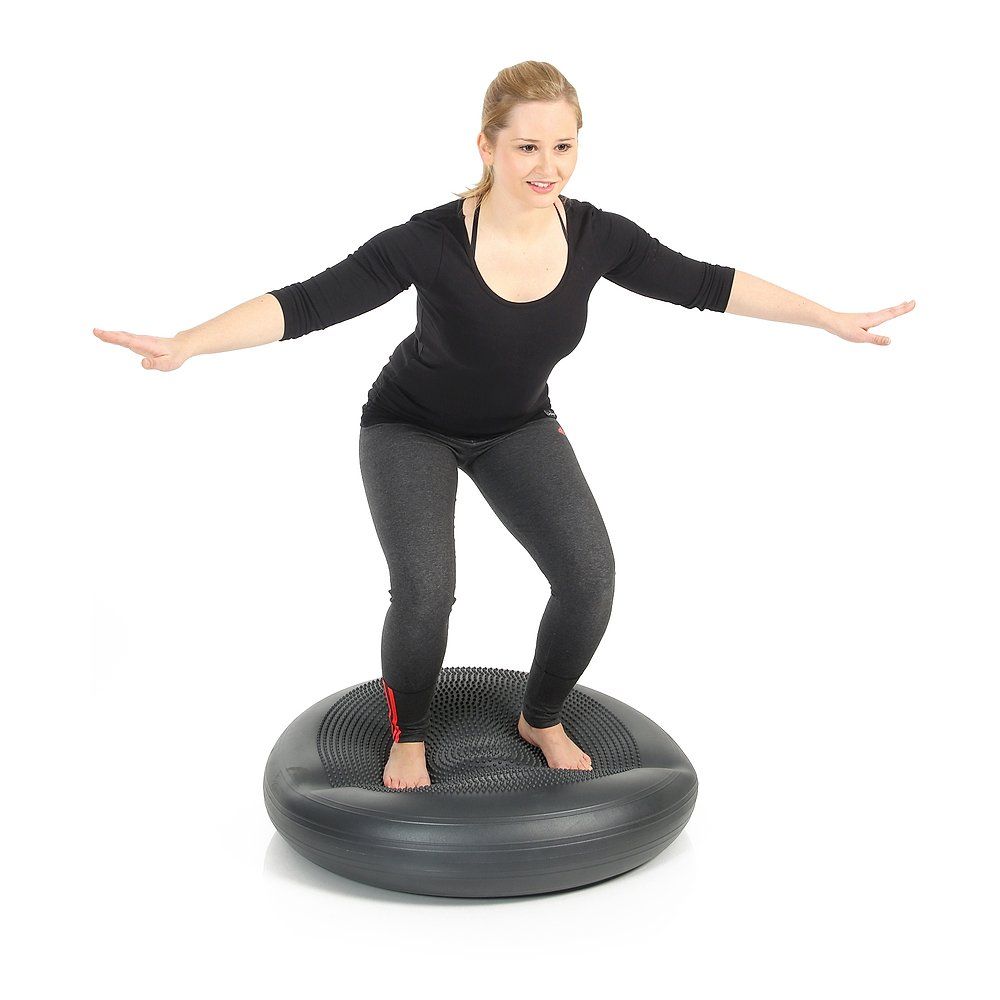 Person steht auf einem grauen, runden Ballkissen. Arme seitlich ausgestreckt. Schwarze Kleidung.