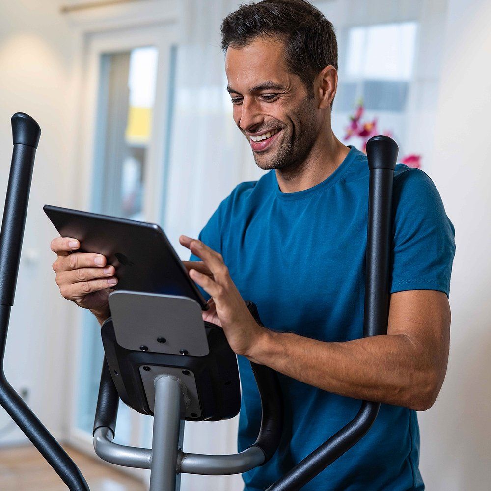 Person benutzt Tablet auf Crosstrainer. Tablet-Halterung. Schwarzes Bedienfeld. Person lächelt. Hintergrund: Raum.