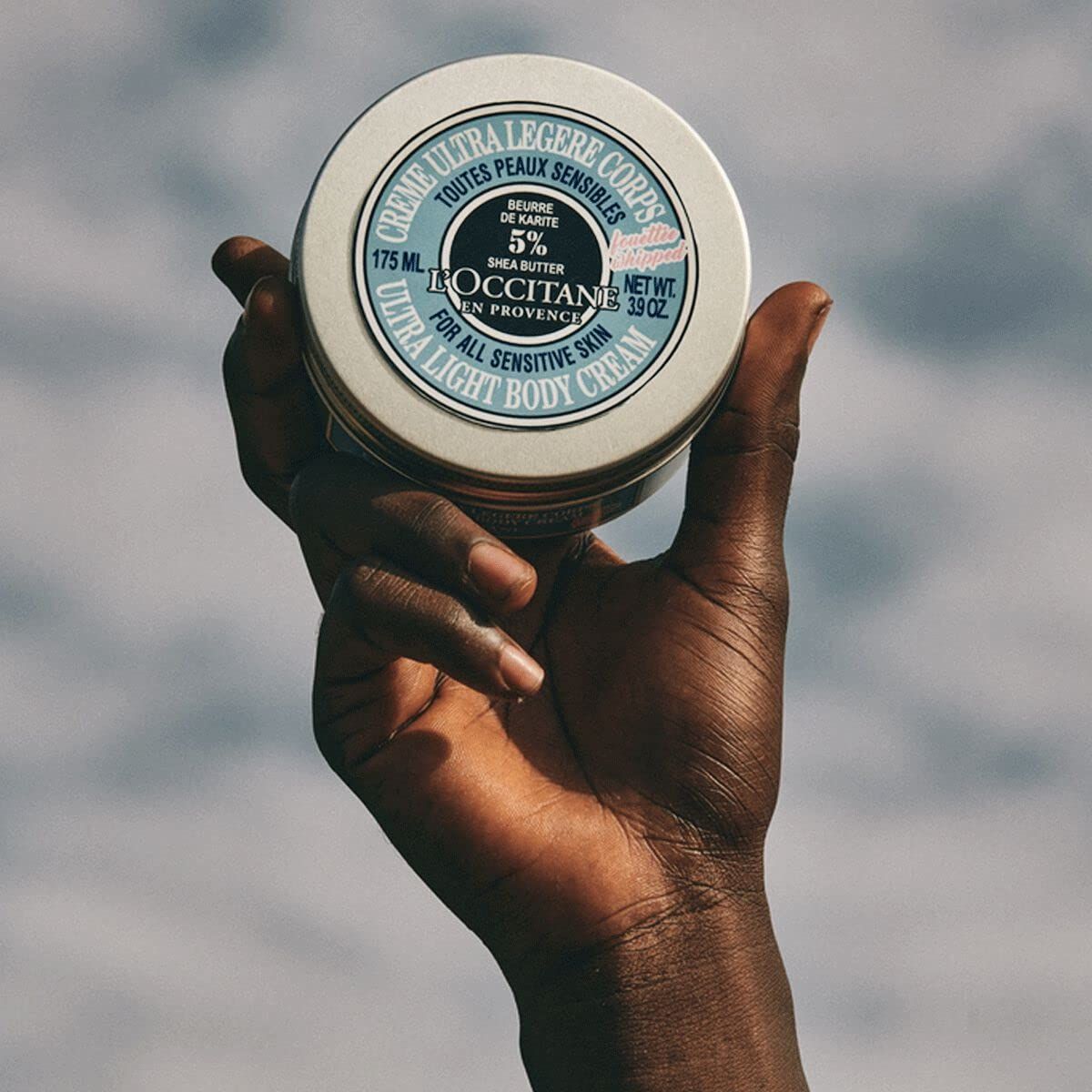 Hand hält eine Dose mit blauem Deckel. Aufschrift: Crème Ultra Légère Corps, L'Occitane, 175 ml, 3.9 oz. Für empfindliche Haut.