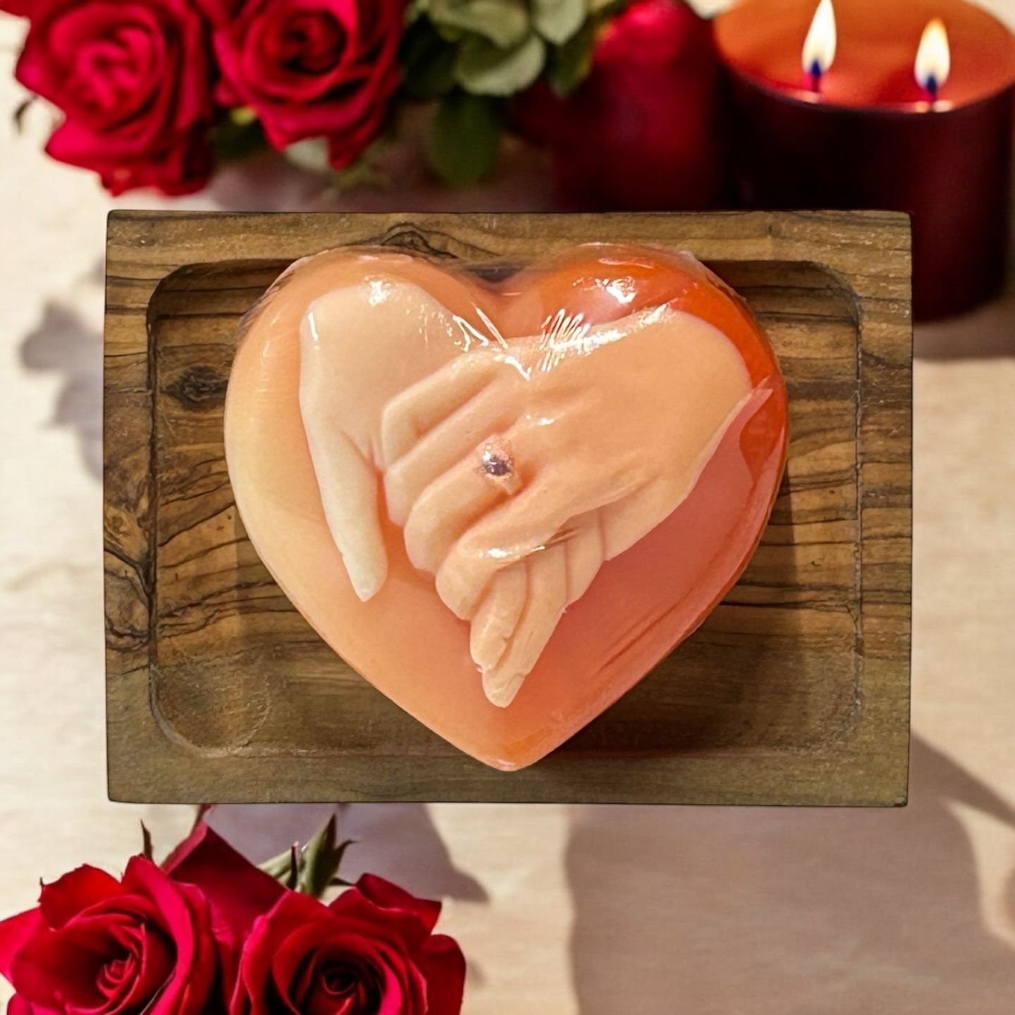 Herzförmige Seife mit Händen, die sich halten. Eine Hand trägt einen Ring. Auf einem Holztablett, umgeben von Rosen und Kerzen.