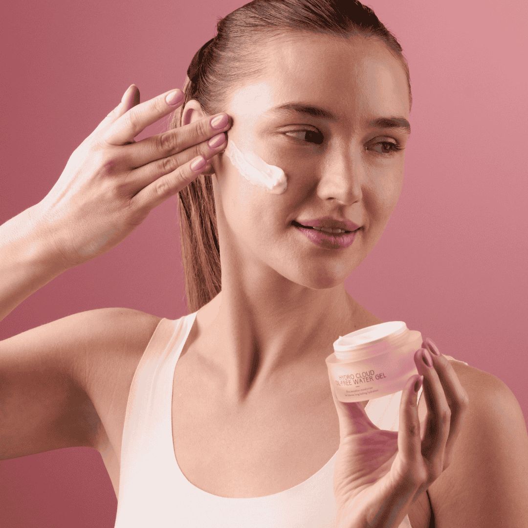 Frau trägt Creme auf das Gesicht auf. Sie hält einen rosa Tiegel mit der Aufschrift HYDRO CLOUD OIL-FREE WATER GEL.