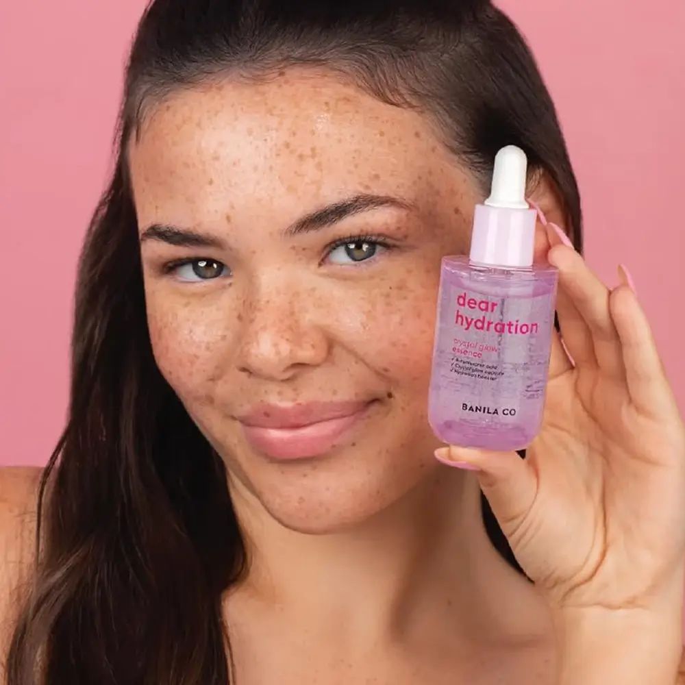 Frau hält eine rosa Serumflasche. Aufschrift: dear hydration, crystal glow essence. Marke: BANILA CO. Hintergrund: rosa.