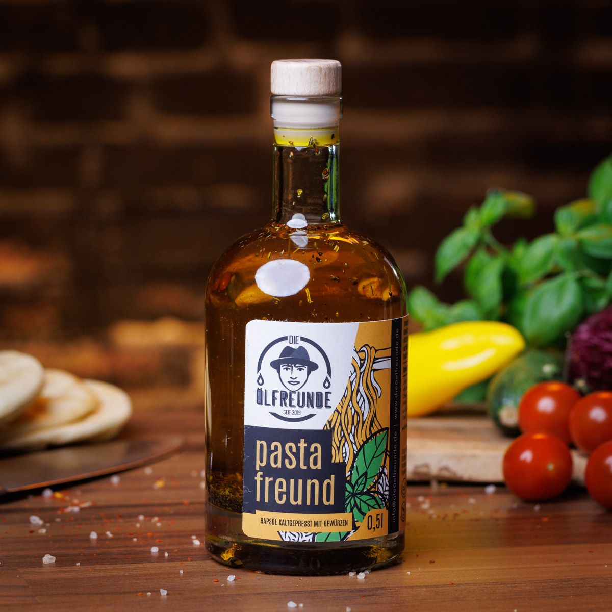 Glasflasche mit gelbem Inhalt und Aufkleber. Aufschrift: Ölfreunde, pasta freund, 0,5l. Mit Holzverschluss. Auf Tisch mit Gemüse.