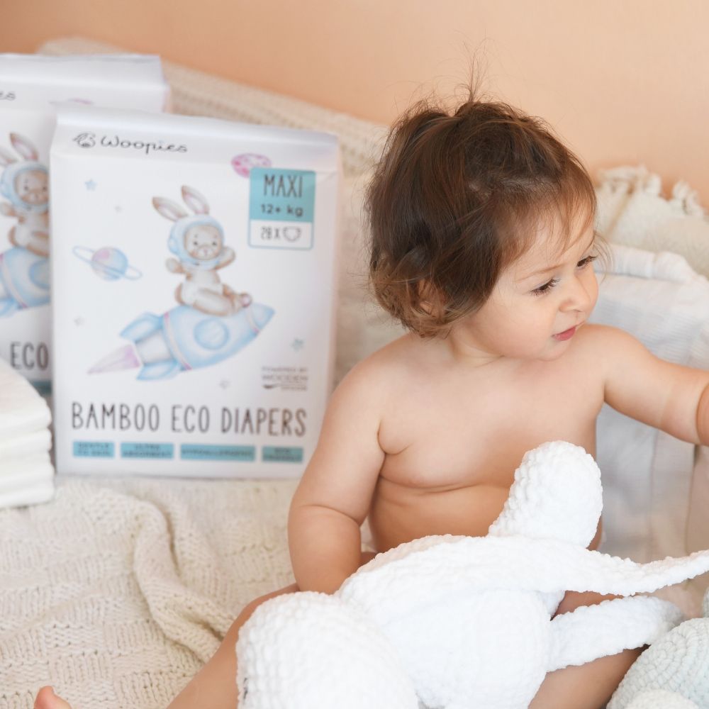 Kleinkind mit Babywindel-Packungen. Aufdruck: Hase auf Rakete, Schriftzug "MAXI 12+ kg", "BAMBOO ECO DIAPERS", "Woopies".