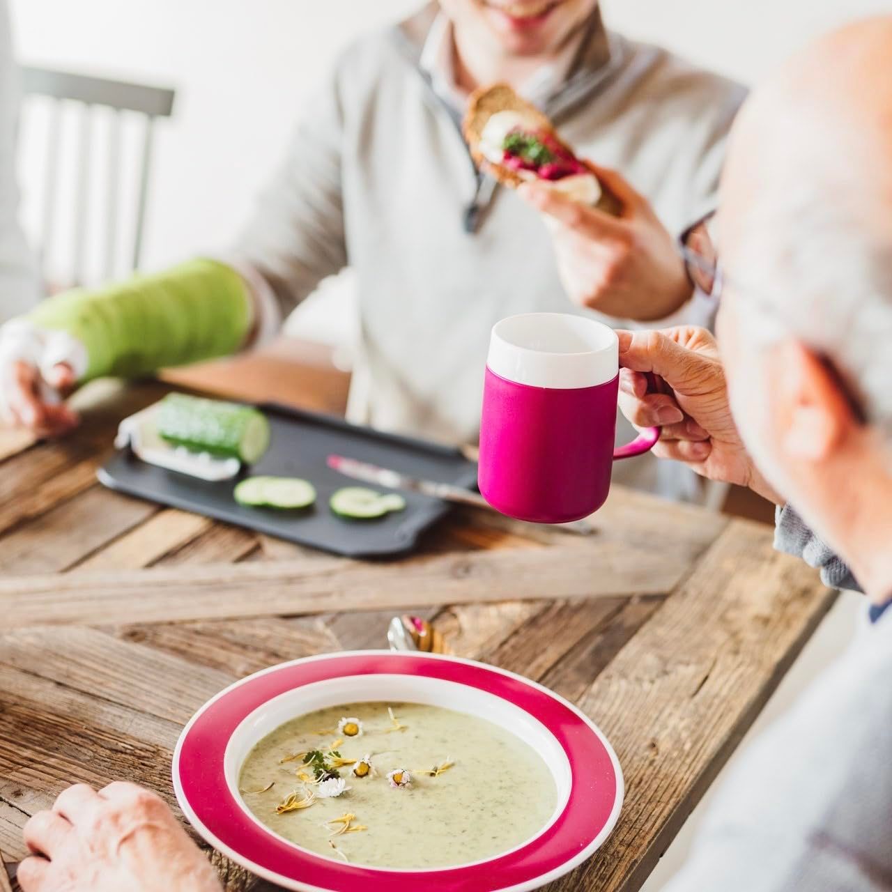 Personen an einem Tisch. Eine Person hält eine pinkfarbene Tasse. Auf dem Tisch Suppe und ein Tablett mit Essen.