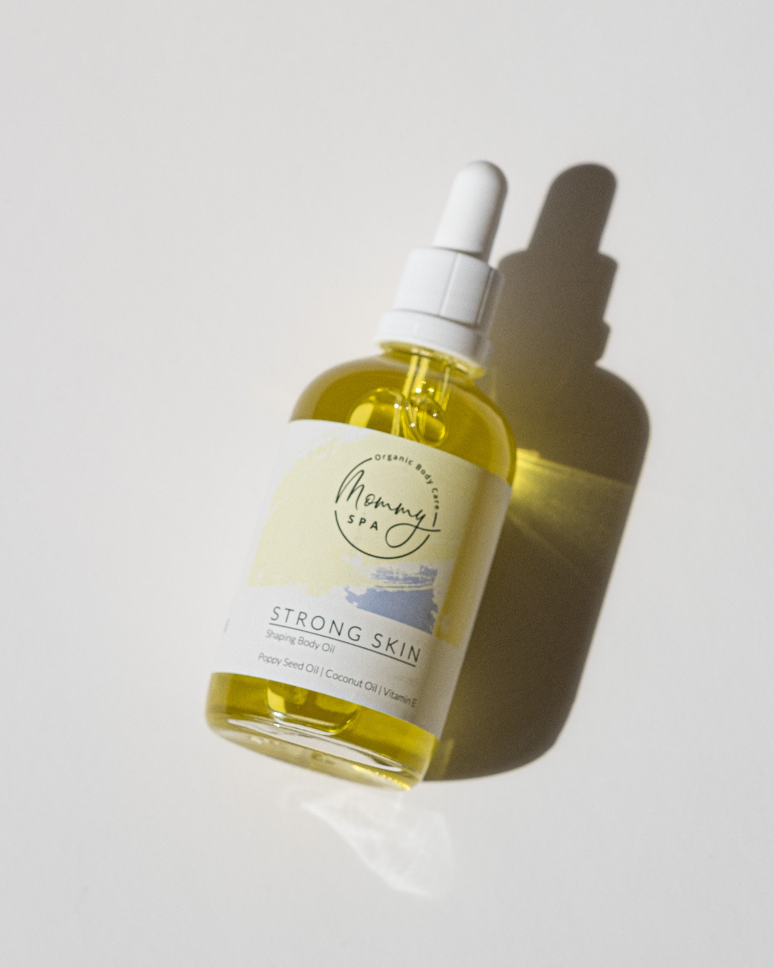 Körperöl-Flasche mit Pipette. Etikett mit "Mommy SPA" und "Strong Skin". Enthält Mohnöl, Kokosöl und Vitamin E. Schatten.