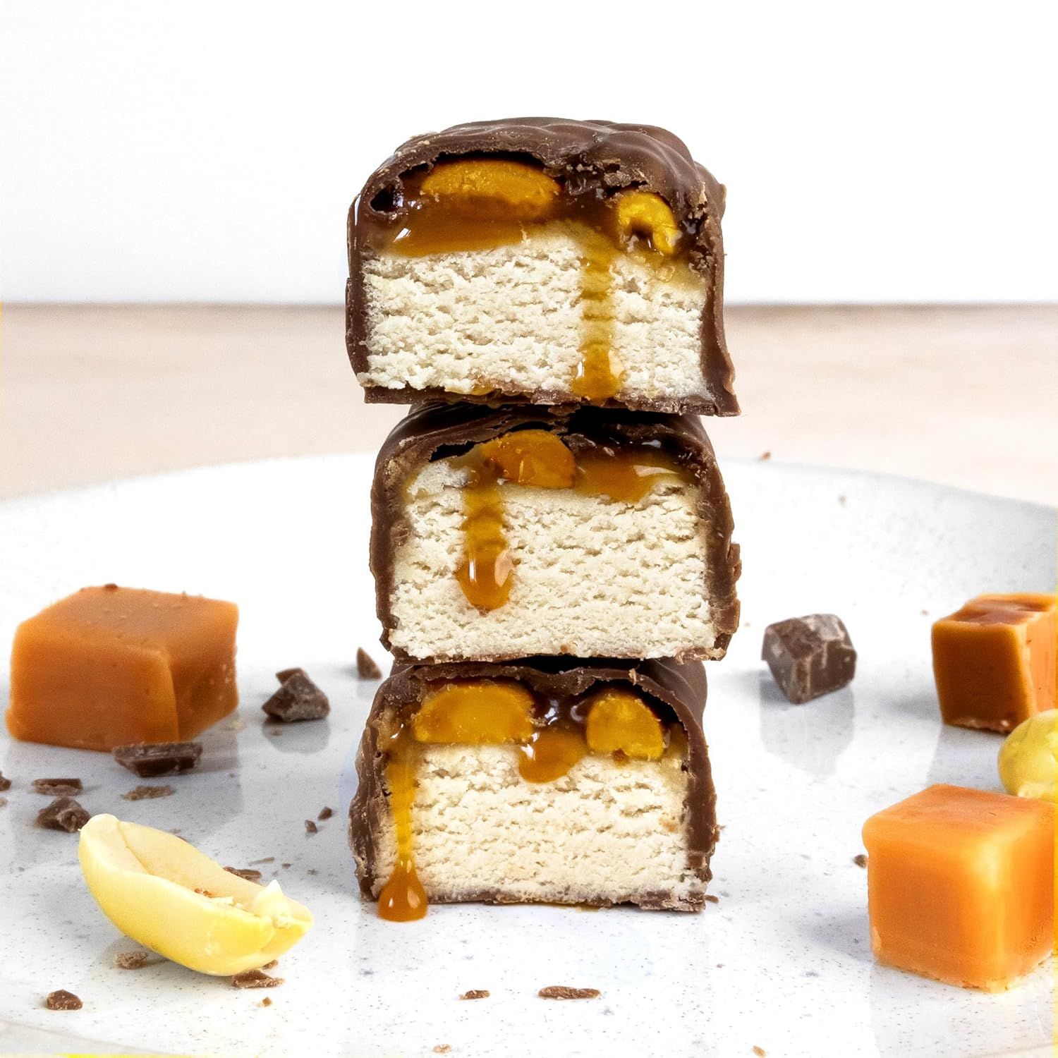 Drei angeschnittene NeoBar-Stücke gestapelt. Schokoladenüberzug, Karamellfüllung, Erdnüsse. Auf einem Teller, umgeben von Karamellstücken.