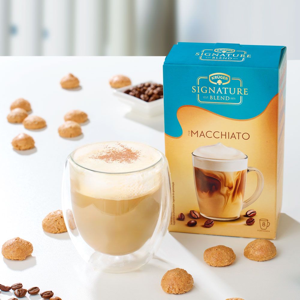 Glas mit Macchiato, daneben Packung Krüger Signature Blend Macchiato. Kekse und Kaffeebohnen.