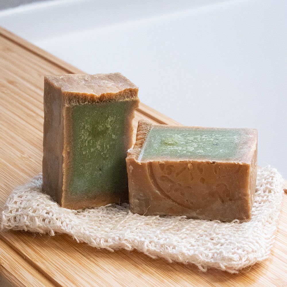 Twee rechthoekige zeepblokken, groene binnenkant. Op een washandje. Op houten plank.
