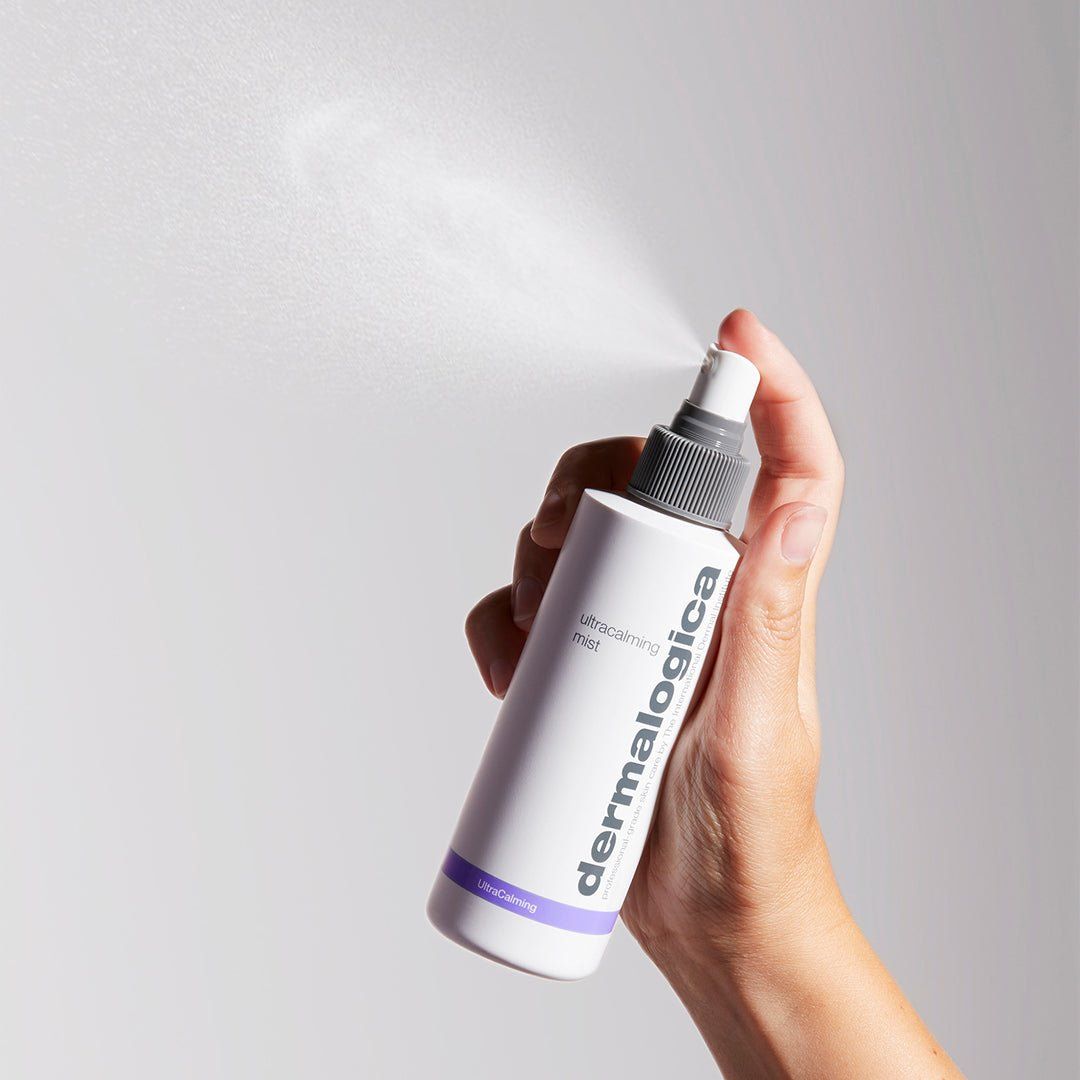 Una mano tiene un flacone di Dermalogica UltraCalming Mist, che spruzza una sottile nebbia. Il flacone è bianco con un erogatore grigio e il logo 'Dermalogica' verticale. 'UltraCalming mist' è scritto sotto. Una striscia viola e 'UltraCalming' sono visibili in basso.