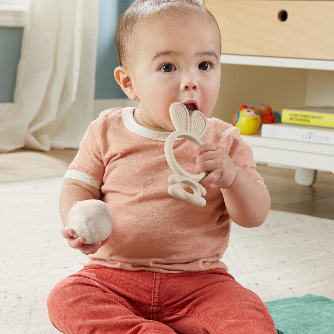 Baby beißt auf einem Hasen-Beißring. Ein Ball liegt in der Hand. Beide Spielzeuge sind hellgrau. Das Baby sitzt auf dem Boden.
