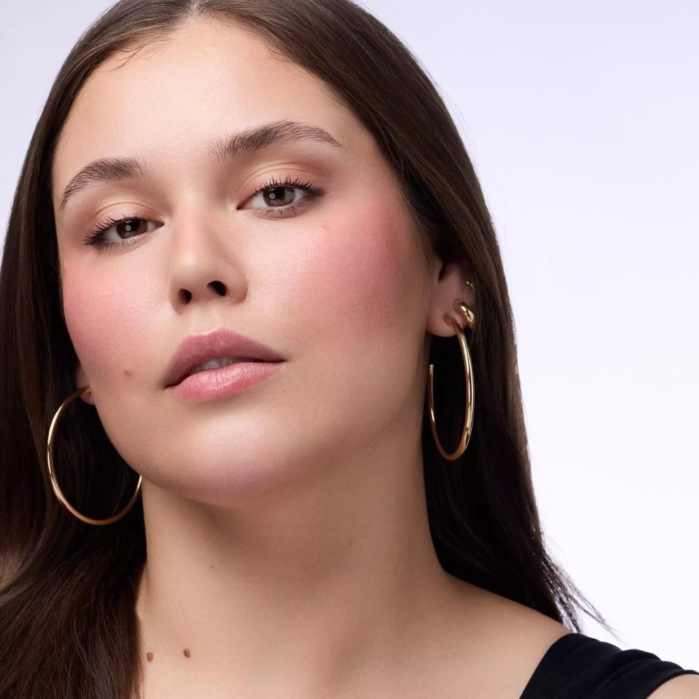 Porträt einer Frau mit goldfarbenen Creolen. Rouge auf den Wangen. Helle Haut, dunkle Haare.