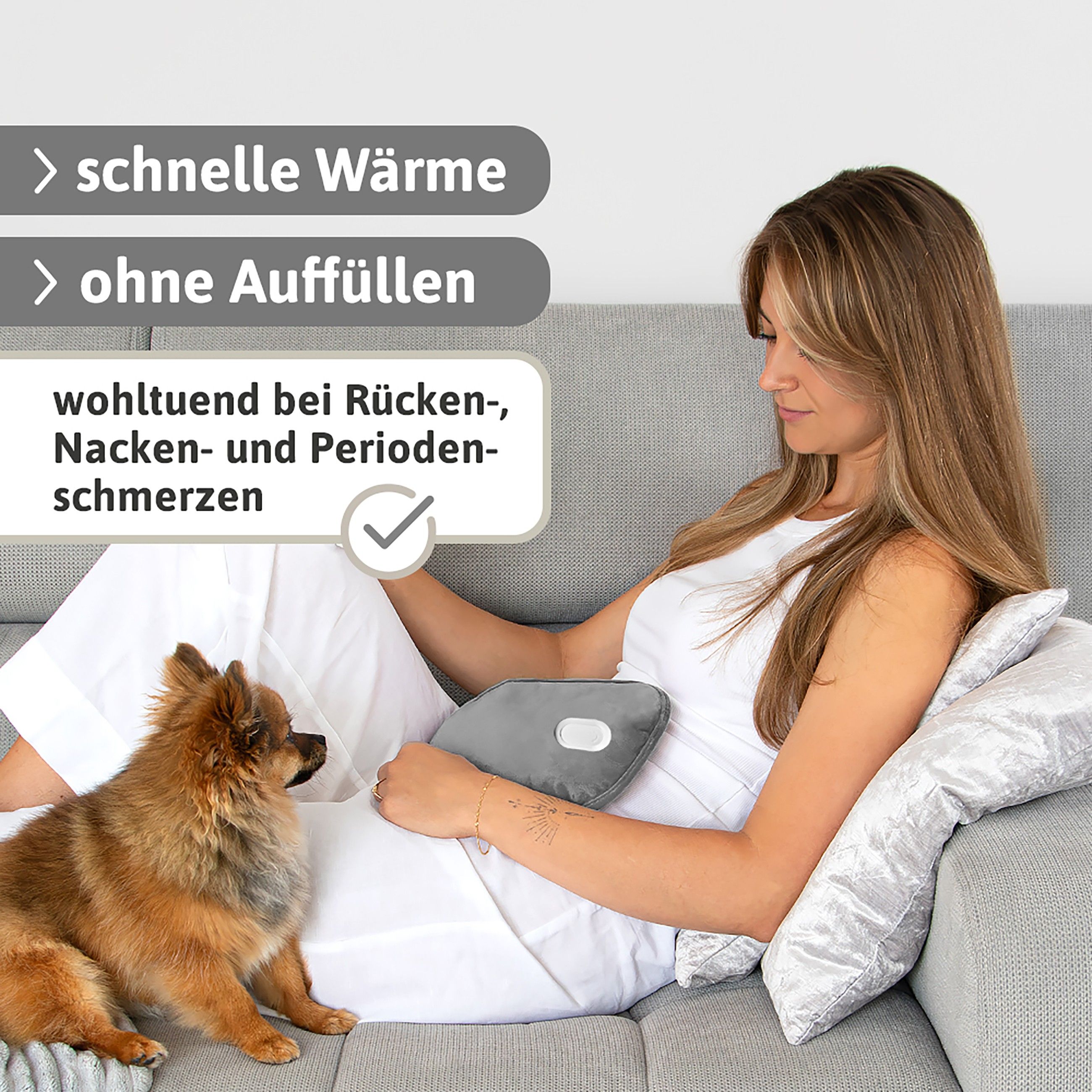 Frau sitzt auf einem Sofa mit einer grauen Wärmflasche. Ein kleiner Hund sitzt neben ihr.