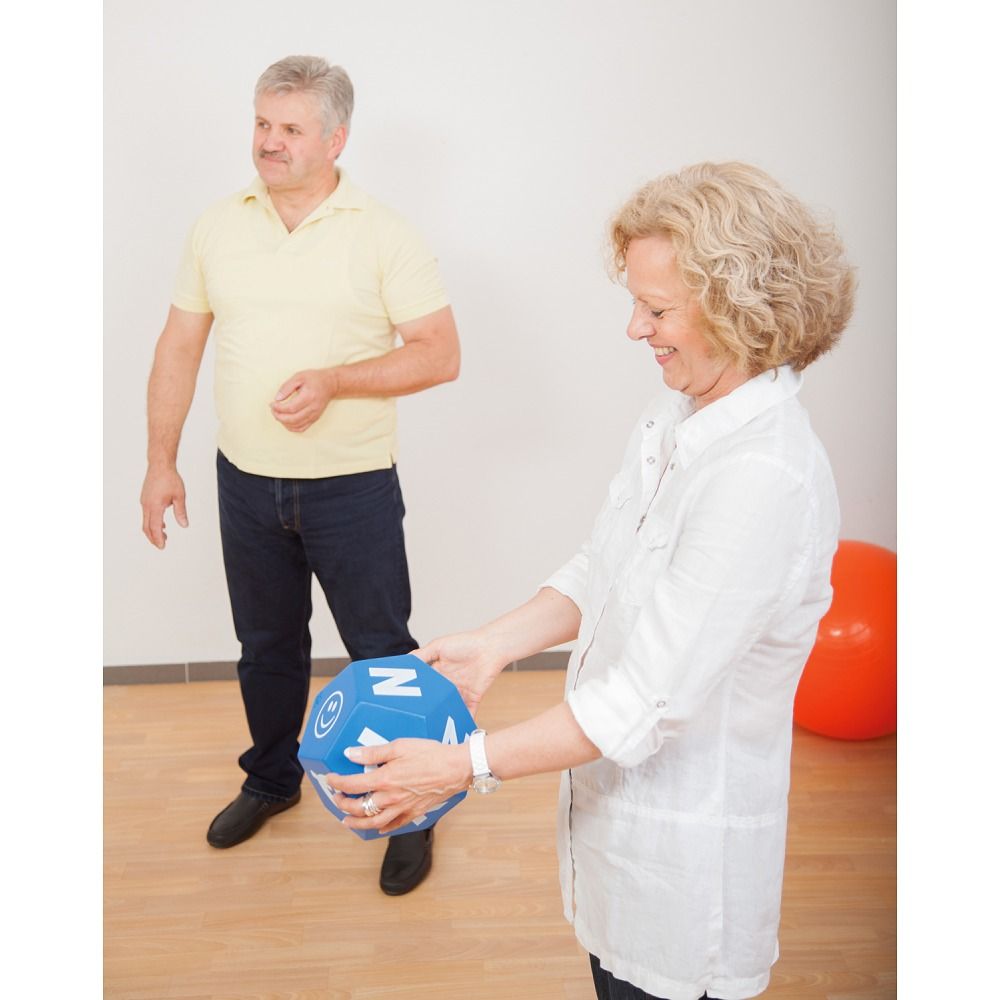 Blauer Schaumstoffwürfel mit Buchstaben. Eine Frau hält den Würfel, ein Mann steht daneben. Gymnastikraum.