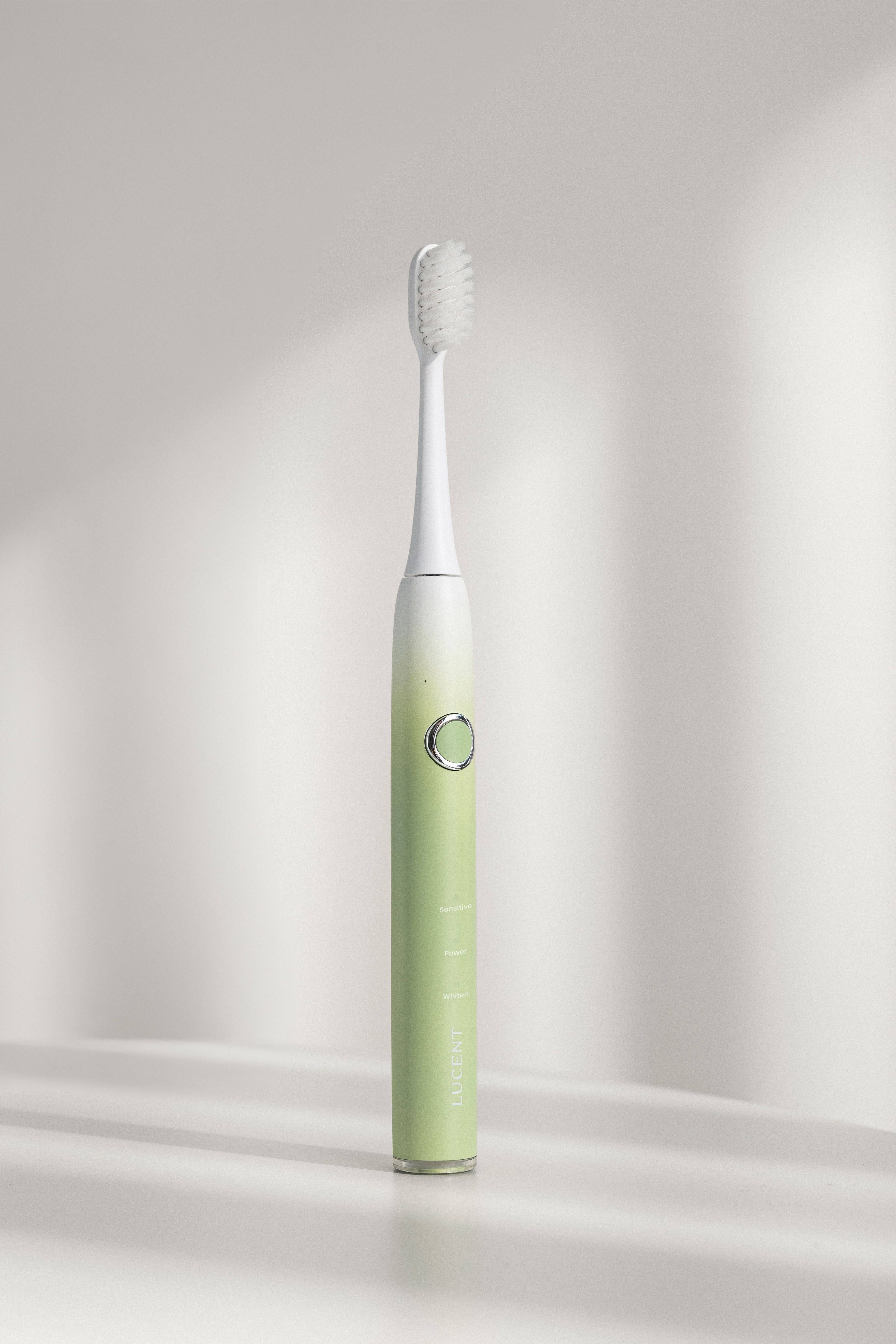 Elektrische Zahnbürste mit grünem Griff und weißem Bürstenkopf. Ein Knopf und Schriftzug sind sichtbar. Schattenwurf.