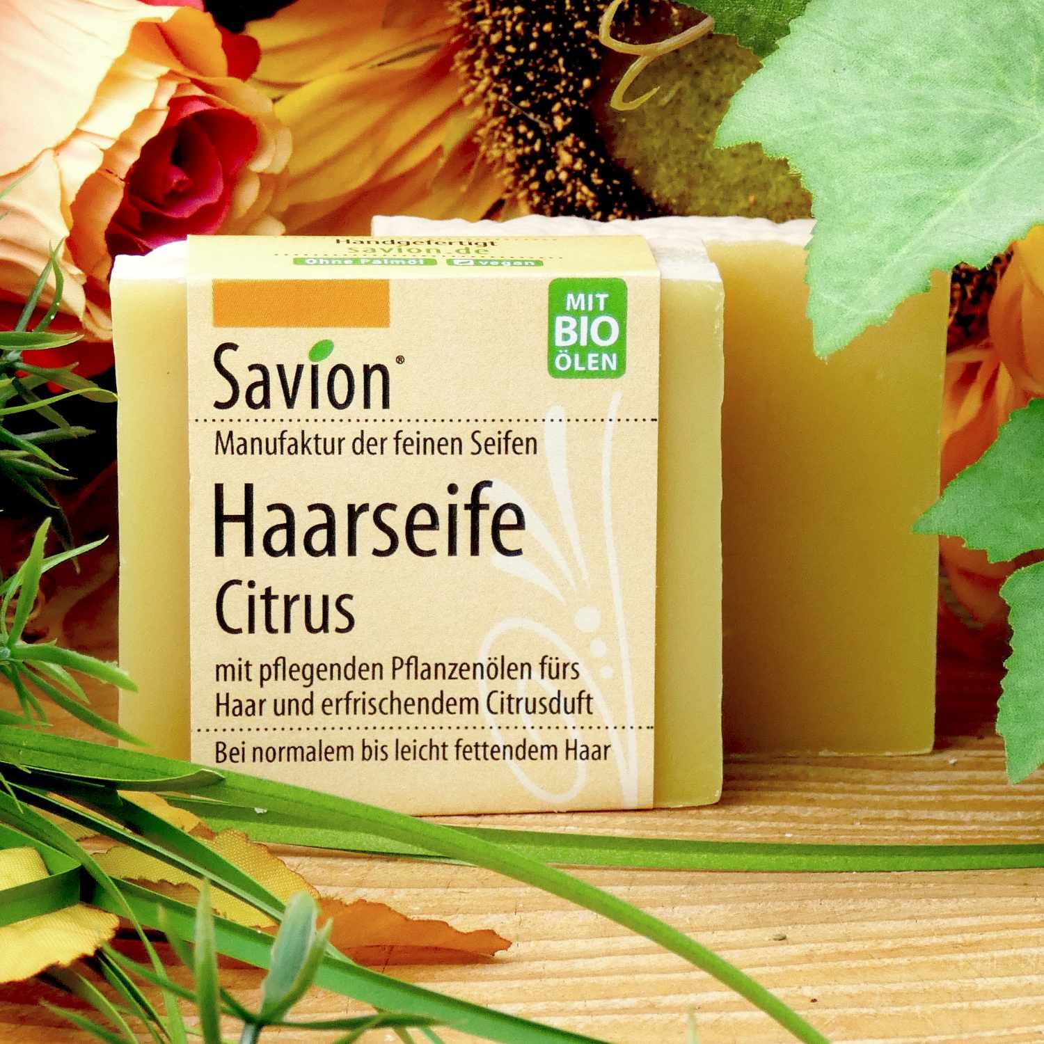 Zwei Seifenstücke mit Papierbanderole. Aufschrift: Savion Manufaktur der feinen Seifen, Haarseife Citrus. Hintergrund mit Blumen.
