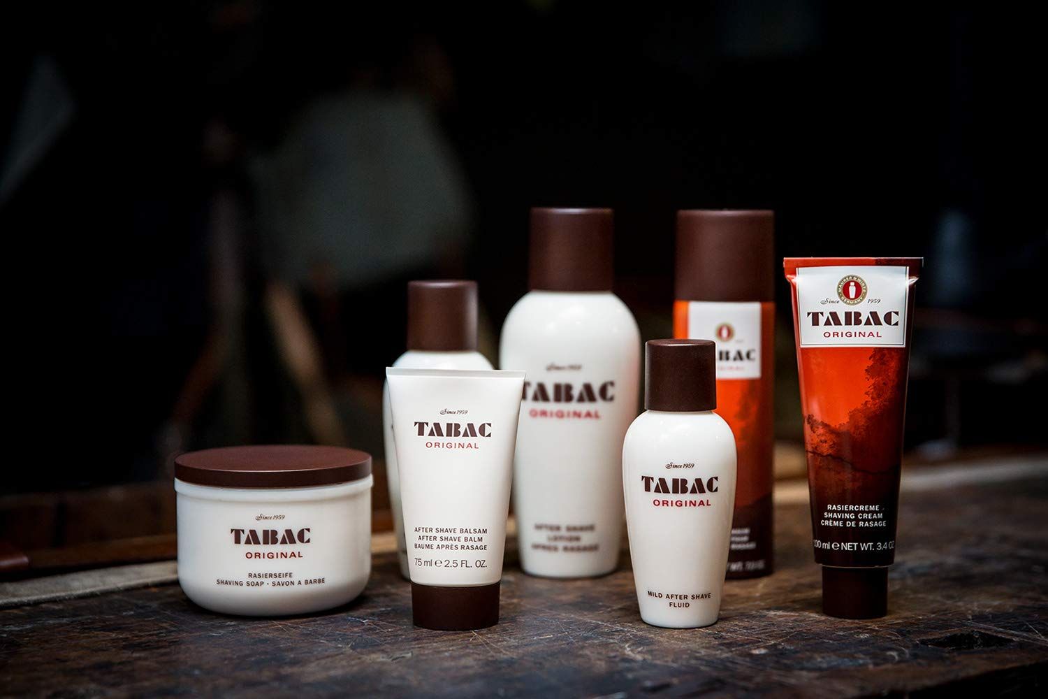 Verschiedene Tabac Original Produkte: Flaschen, Tiegel, Tuben. Braune Deckel und Schriftzug. Auf dunklem Hintergrund.
