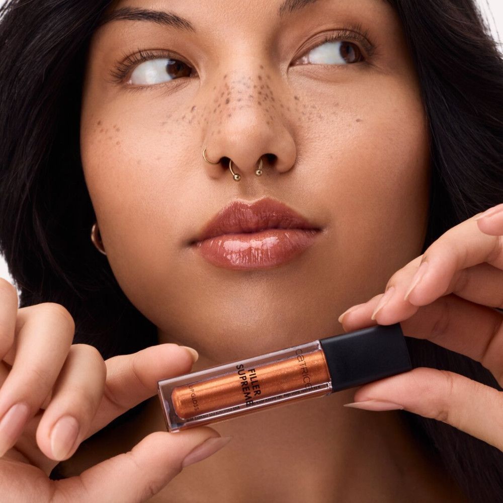 Person hält Catrice Supreme Lippenfiller Gloss. Produkt in transparenter Tube mit braunem Gloss. Schwarzer Deckel.