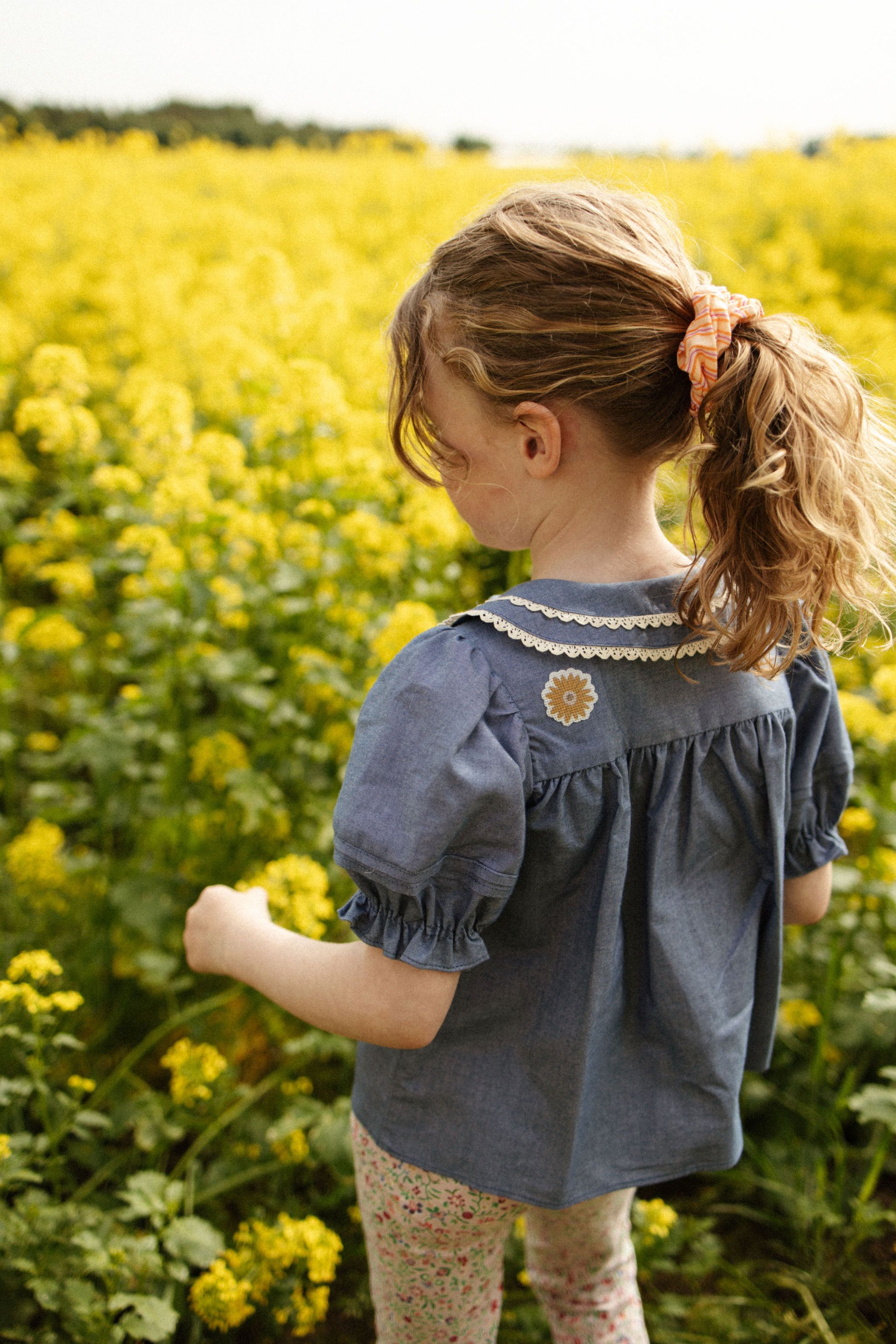 Kind in blauem Hemd mit Blumen-Applikation. Steht in einem Feld mit gelben Blumen.