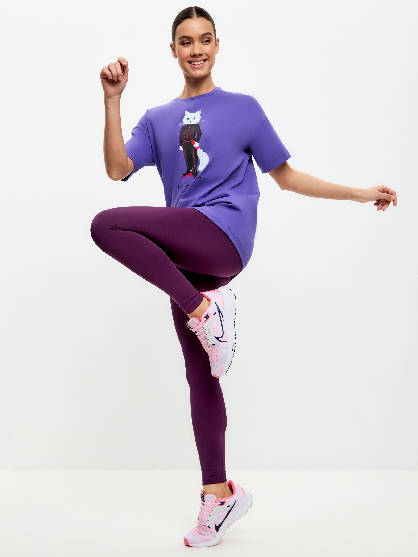 Dunkelviolette Leggings. Frau trägt lila T-Shirt mit Katzenmotiv. Pinke Schuhe. Pose mit erhobenem Bein.