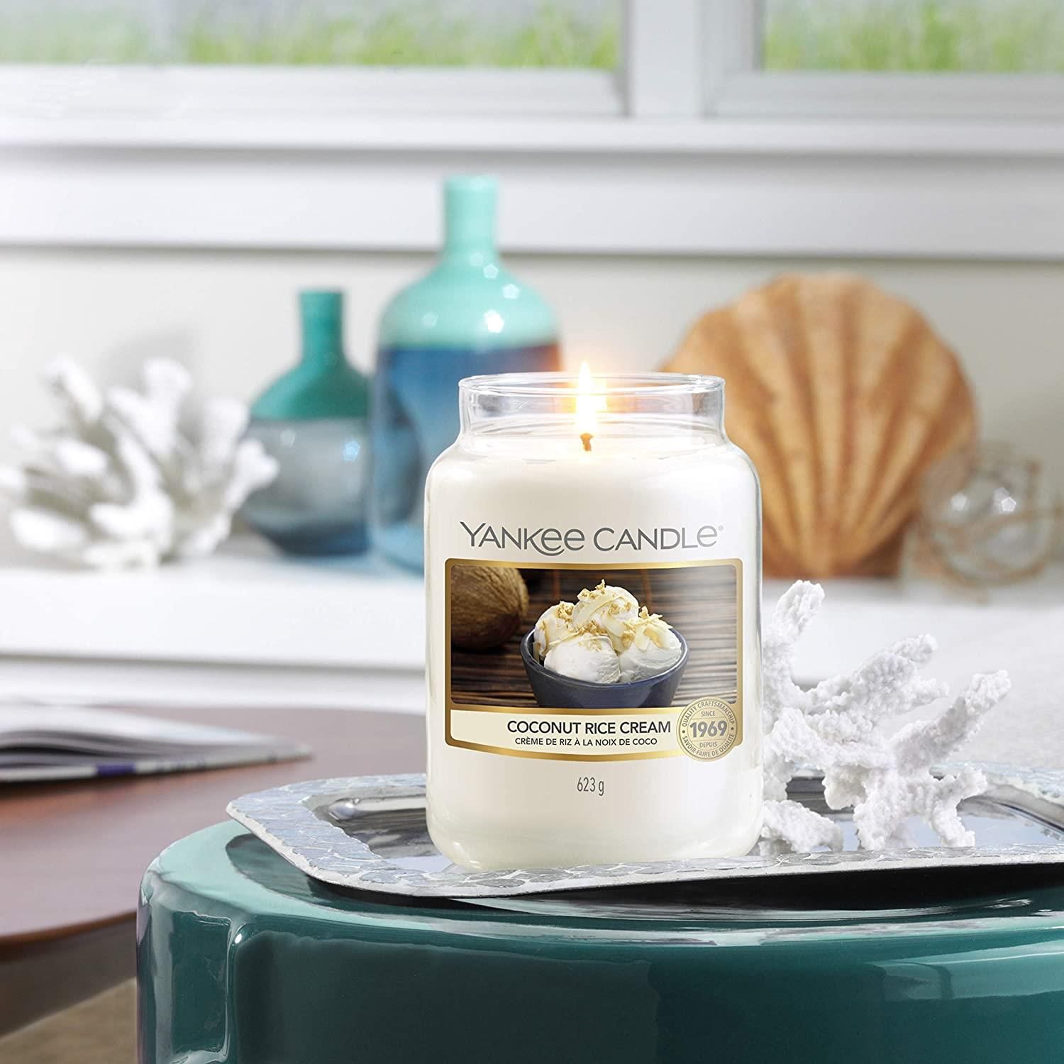 Glas-Duftkerze mit Deckel, brennend. Aufkleber mit Produktbild und Schriftzug: Coconut Rice Cream. Marke: Yankee Candle. 623g.