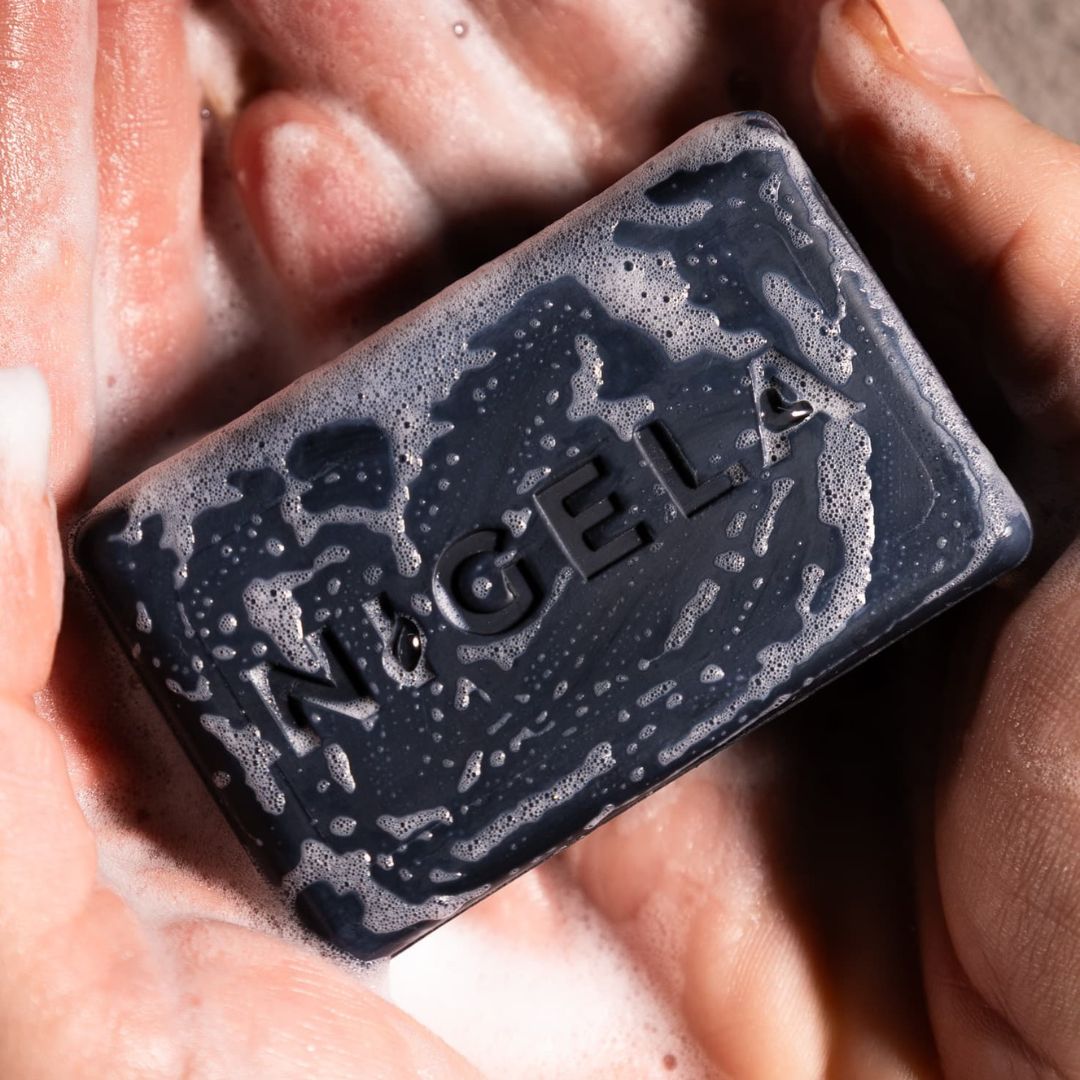 Schwarze Seife mit Nigela-Logo, von Händen gehalten und mit Schaum bedeckt.
