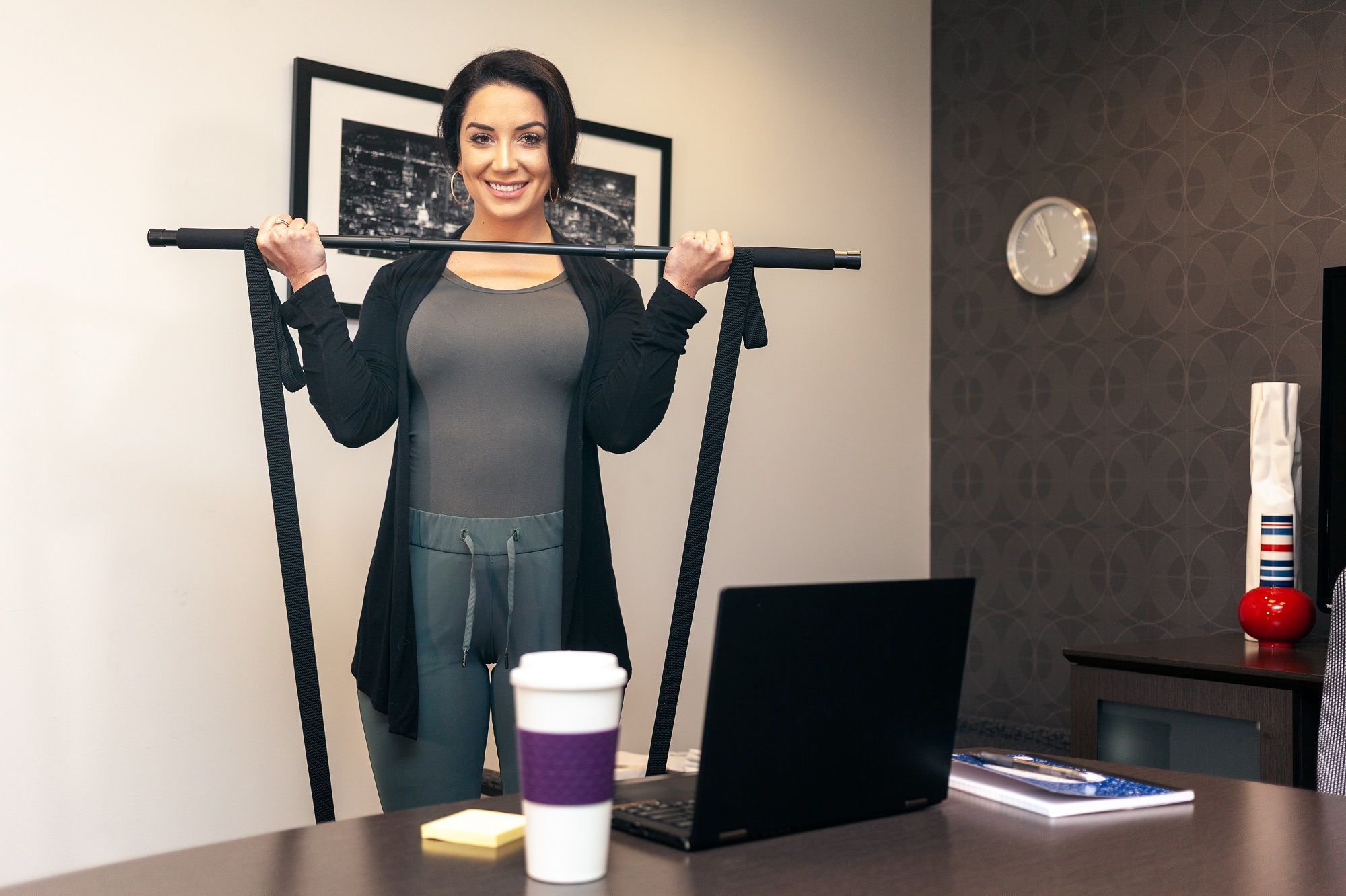 Frau mit Trainingsgerät in einem Büro. Schwarze Stange, Laptop, Kaffeetasse. Graues Oberteil, schwarze Jacke.