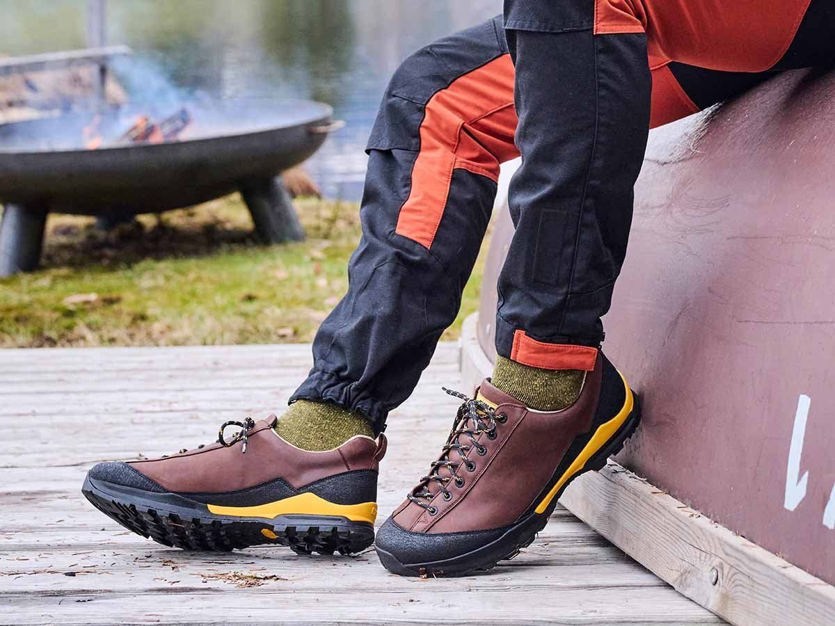 Person trägt braune Wanderschuhe mit gelben Akzenten. Schwarze Hose und grüne Socken. Feuer im Hintergrund.