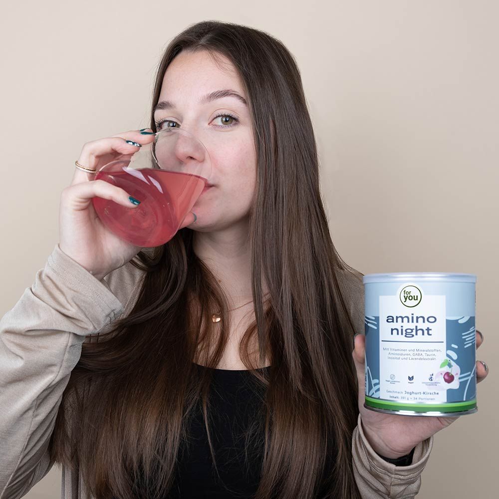 Frau trinkt aus Glas mit roter Flüssigkeit, neben Dose "for you amino night". Produktetikett sichtbar. Frau mit langen Haaren, neutraler Hintergrund.