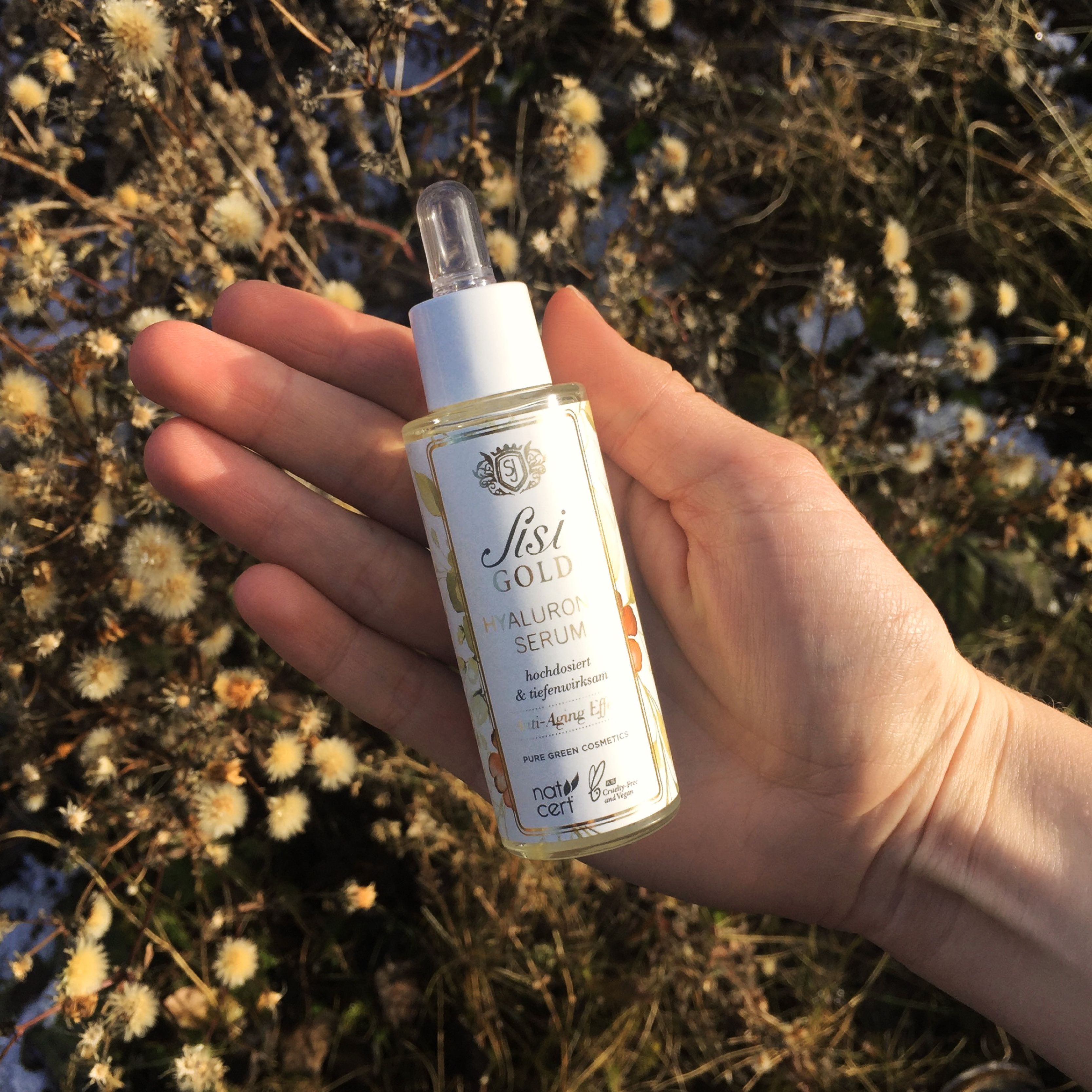 Serumflasche mit Pipette, gehalten in einer Hand. Aufschrift: Sisi GOLD Hyaluronic Serum. Hintergrund: Pflanzen.