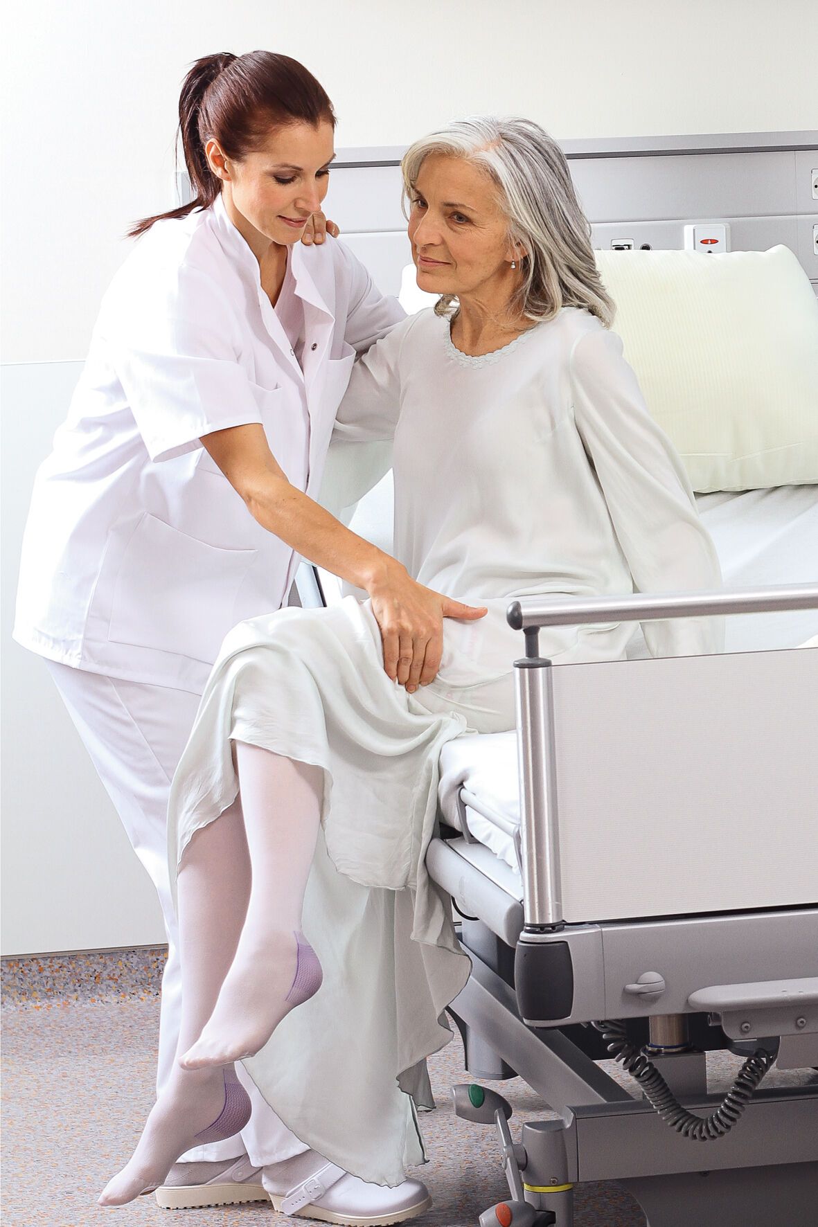 Eine ältere Frau sitzt auf einem Bett und wird von einer Krankenschwester unterstützt. Sie trägt weiße Kompressionsstrümpfe. Die Krankenschwester trägt eine weiße Uniform. Der Hintergrund ist ein Krankenhauszimmer.