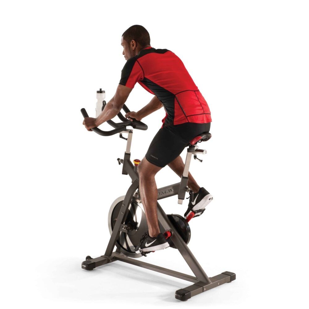Indoor Cycle. Grauer Rahmen, schwarze Lenkstange, roter Sattel. Person trainiert. Wasserflasche am Lenker.