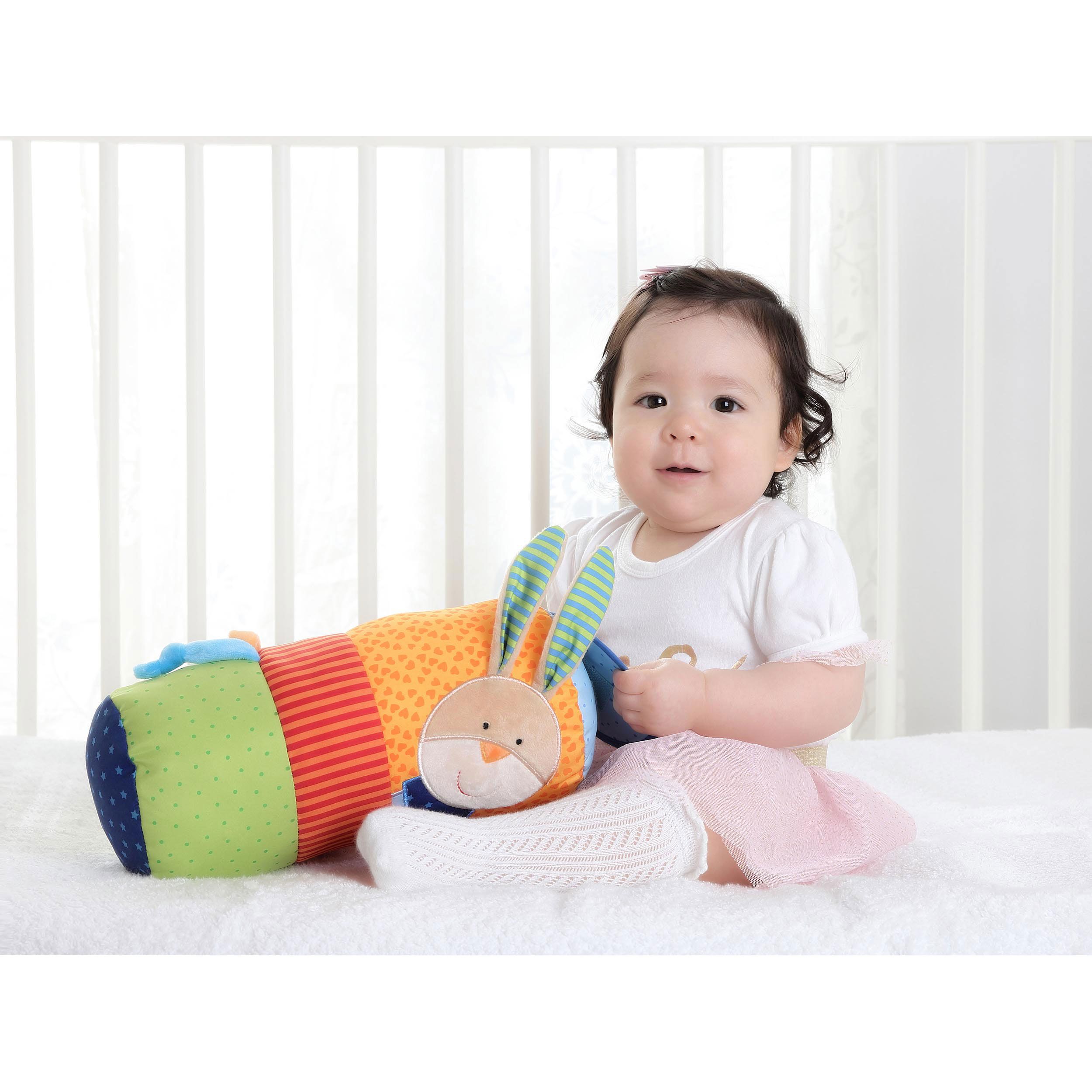 Baby sitzt mit Spielzeug. Zylindrisches Spielzeug mit Hasen-Gesicht. Mehrfarbig. Baby trägt Rock und Strümpfe.